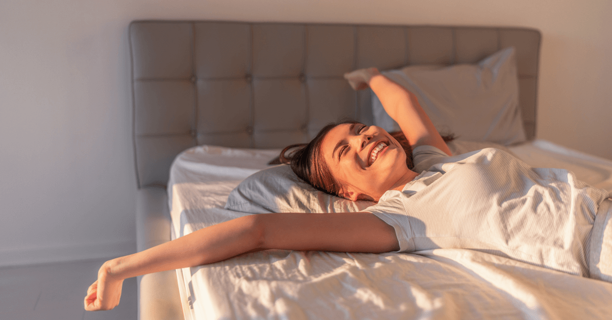 Woman waking up in bed with her arms stretched out, smiling and feeling refreshed after a restful night's sleep