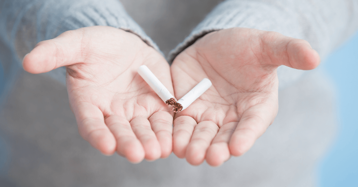 A person wearing a light grey sweater holds the two halves of a broken cigarette in their open palms. The soft lighting and blurred background highlight the act of breaking free from the habit.