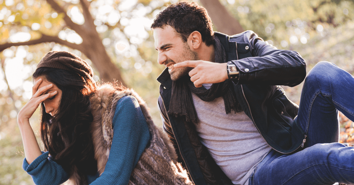 A young man with dark, curly hair and stubble, wearing a black leather jacket and scarf, appears to be arguing with a woman outdoors in a park-like setting with autumn foliage. He is pointing aggressively and leaning towards her with an intense expression.