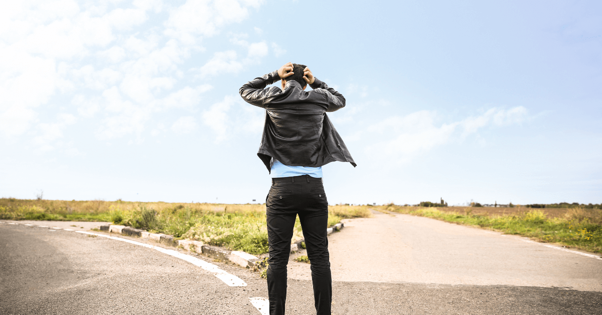 A man dressed in a black leather jacket and dark jeans stands at a fork in the road, holding his head in his hands as he faces a difficult decision.